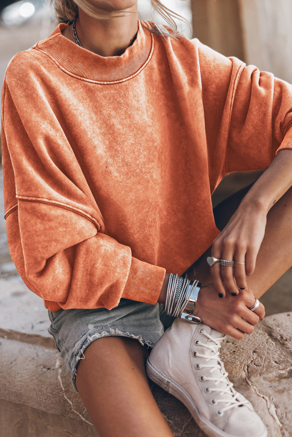 Person wearing an orange sweatshirt and denim shorts sitting on a stone surface.