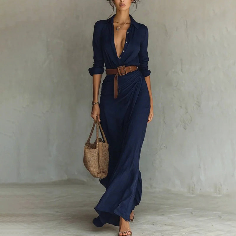 Woman wearing a long navy dress with a belt, holding a woven bag against a plain background