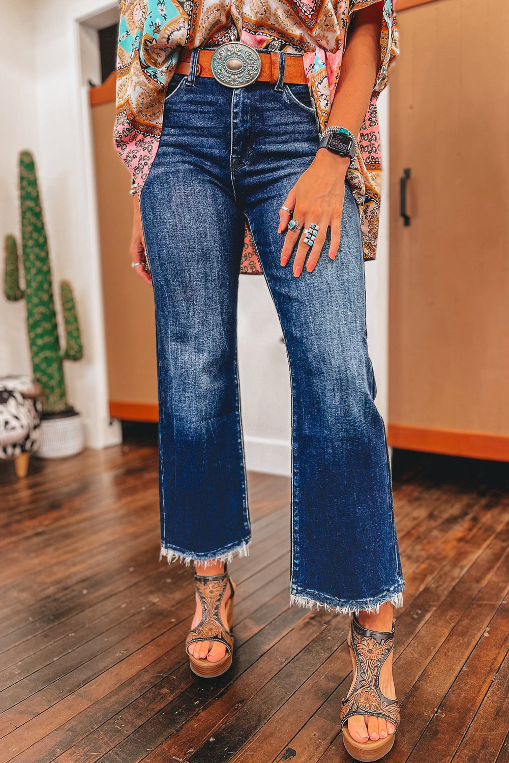 Person wearing blue jeans with a patterned top and sandals on a wooden floor.