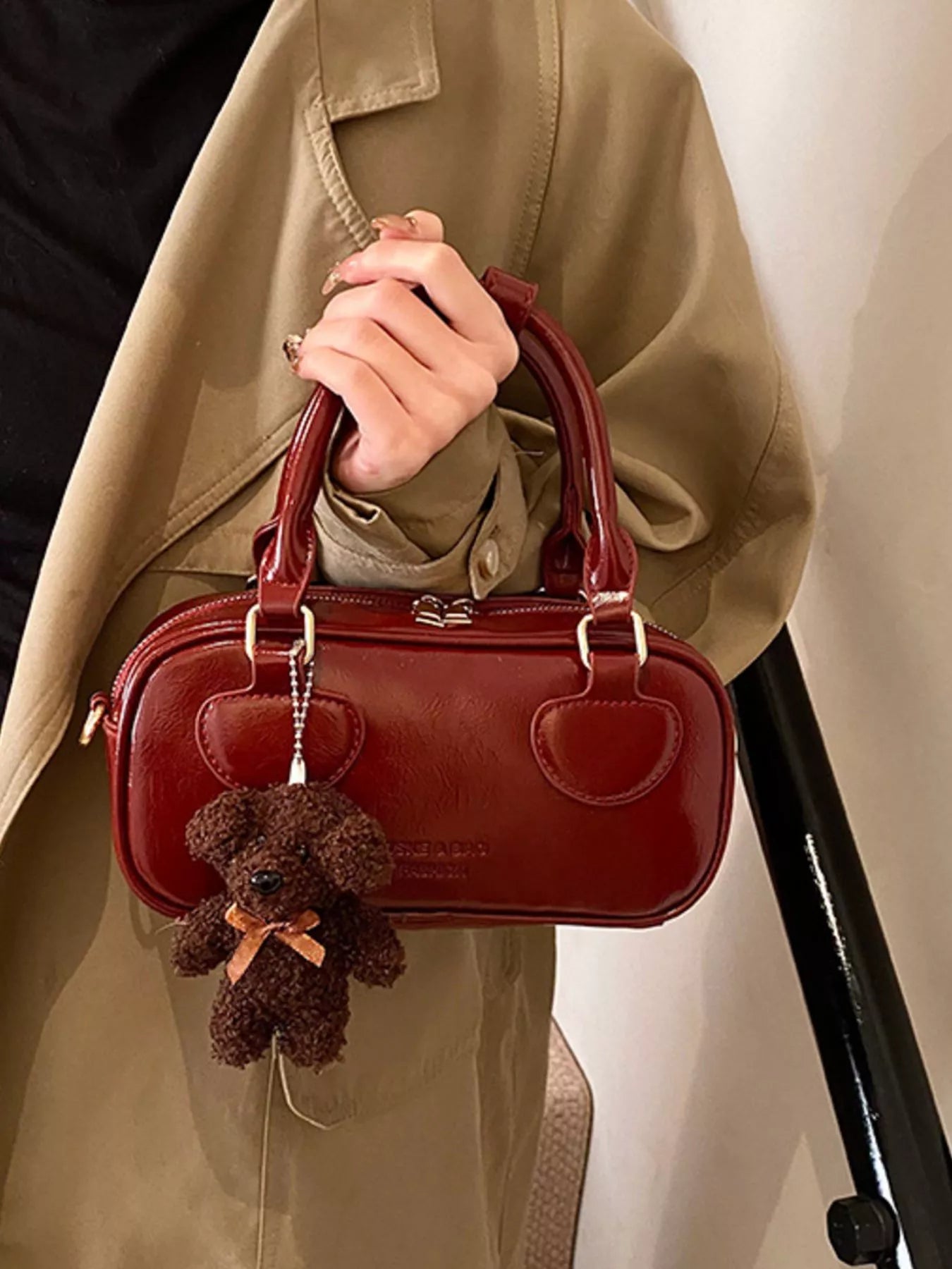Red handbag with a small brown teddy bear charm held by a person wearing a beige coat.