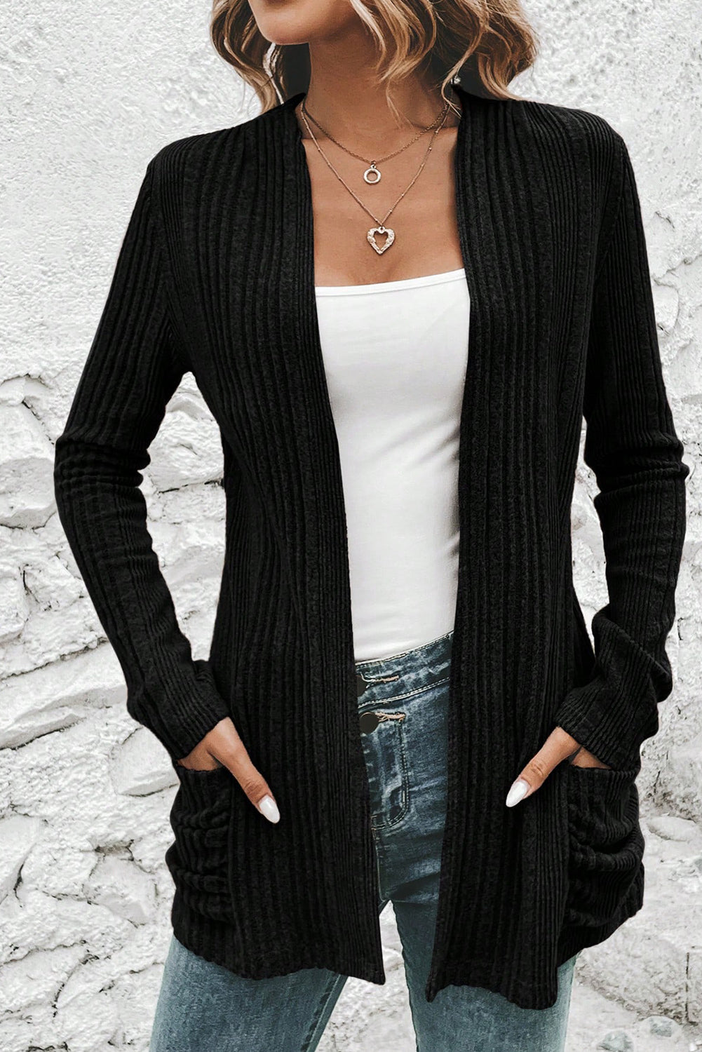 Person wearing a black ribbed cardigan over a white top and blue jeans against a stone wall background.