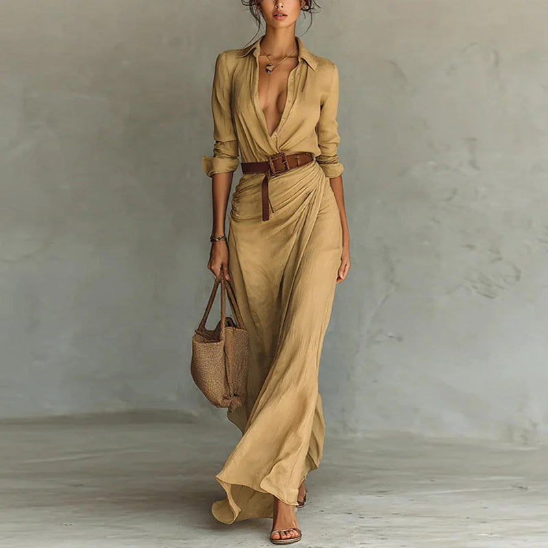 Woman wearing a long beige dress with a belt, holding a woven bag against a plain background