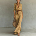 Woman wearing a long beige dress with a belt, holding a woven bag against a plain background