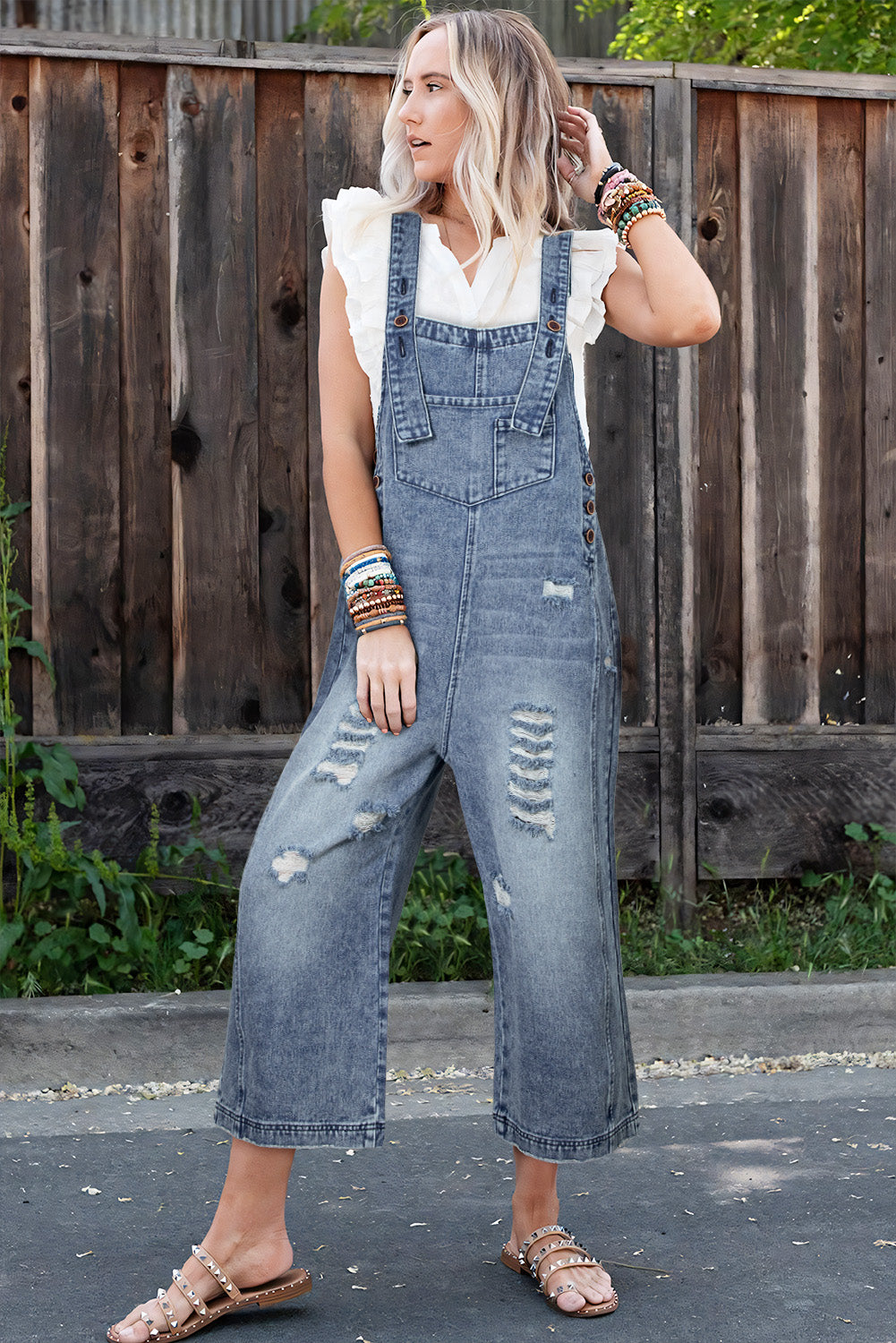 Woman wearing denim overalls standing in front of a wooden fence.