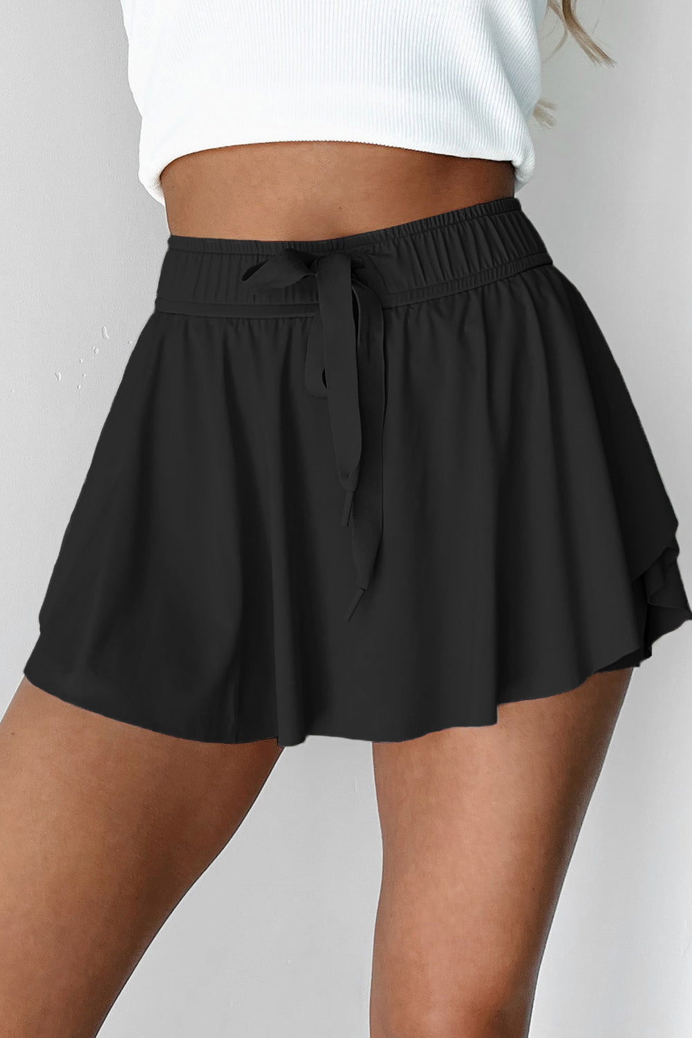 Black tennis skirt with tie waist worn by a person on a plain background