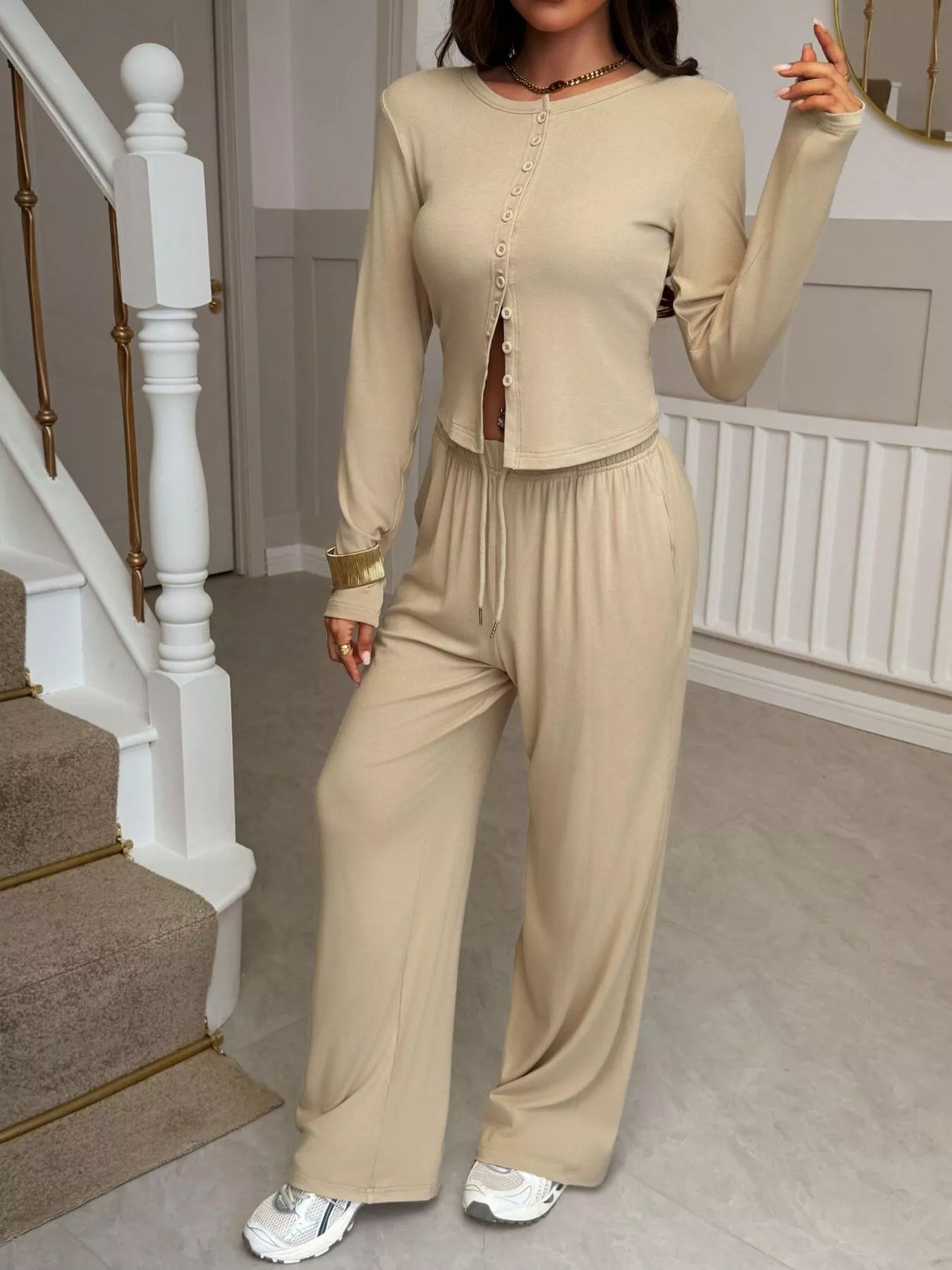 Person wearing a beige outfit standing in a home setting with a staircase.