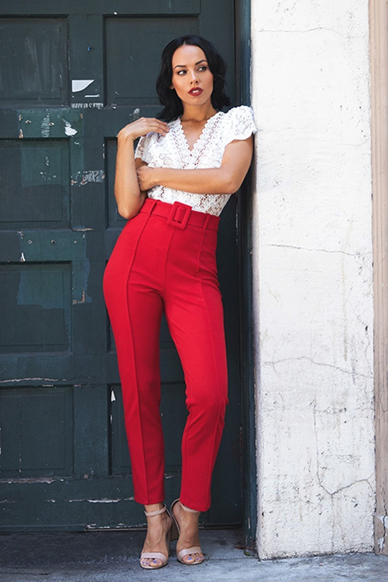 Woman in a white top and red pants leaning against a wall.