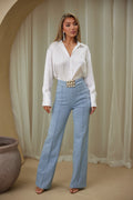 Woman wearing a white blouse and light blue pants standing against a beige curtain.
