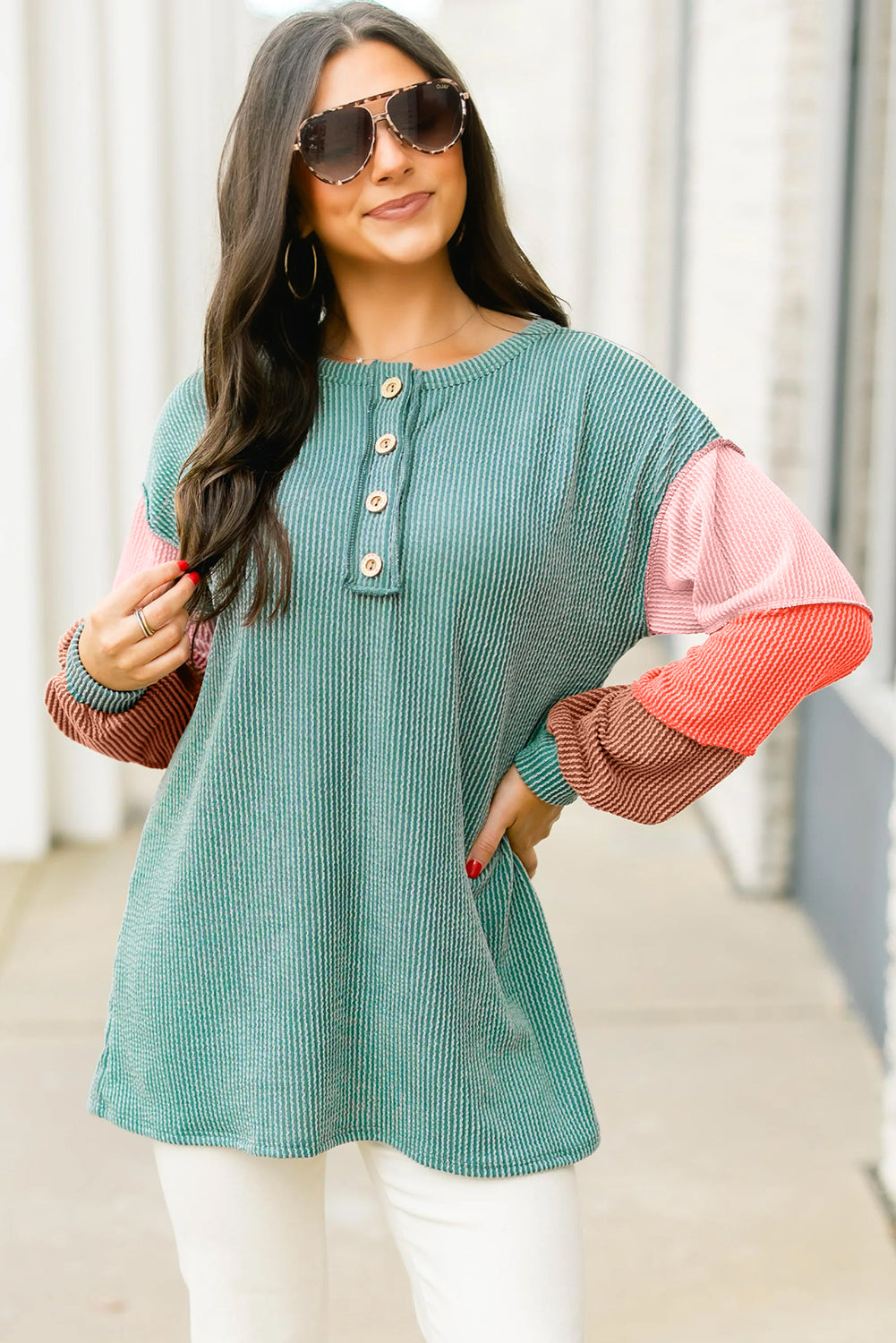 Woman wearing a teal and pink top with sunglasses, standing against a light background.