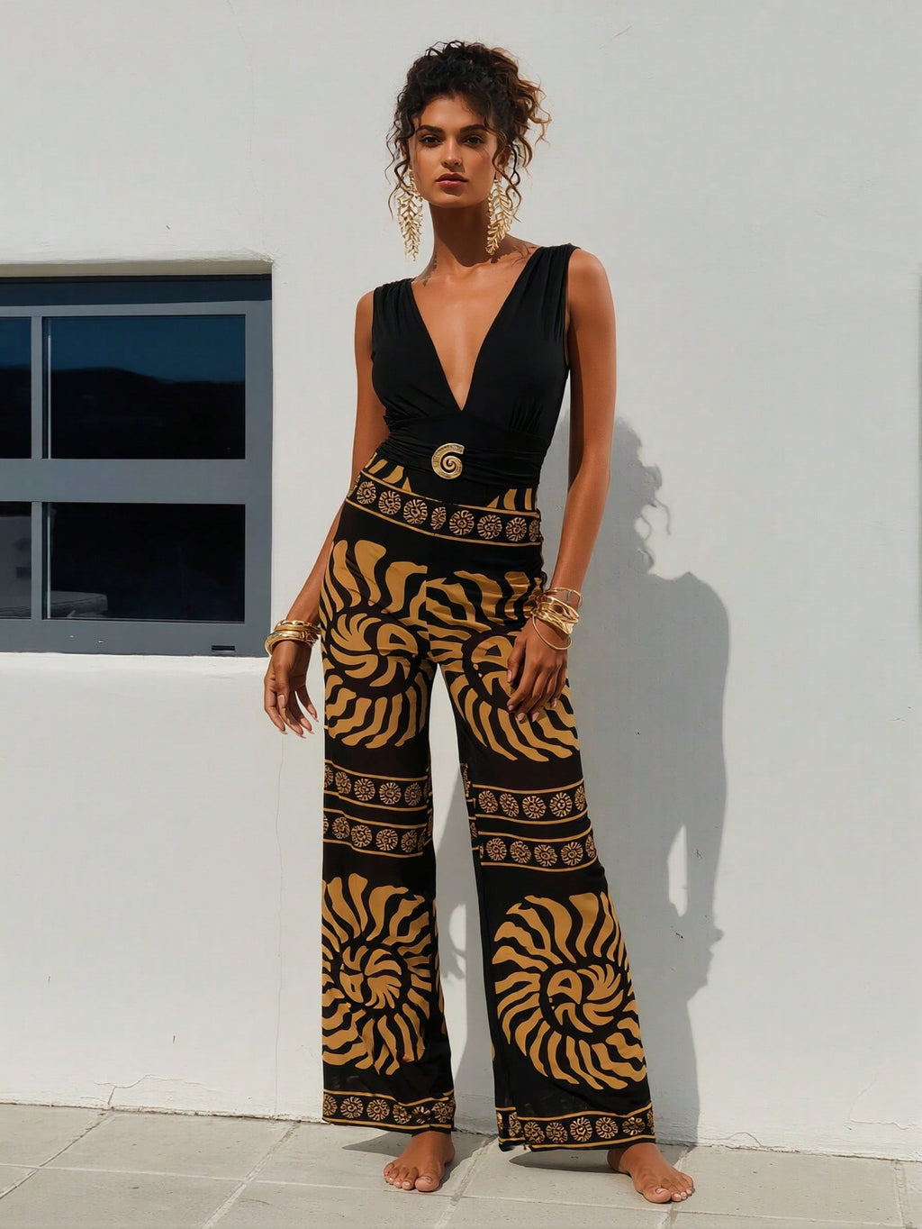 Woman wearing a black and gold patterned jumpsuit standing against a white wall. Front view