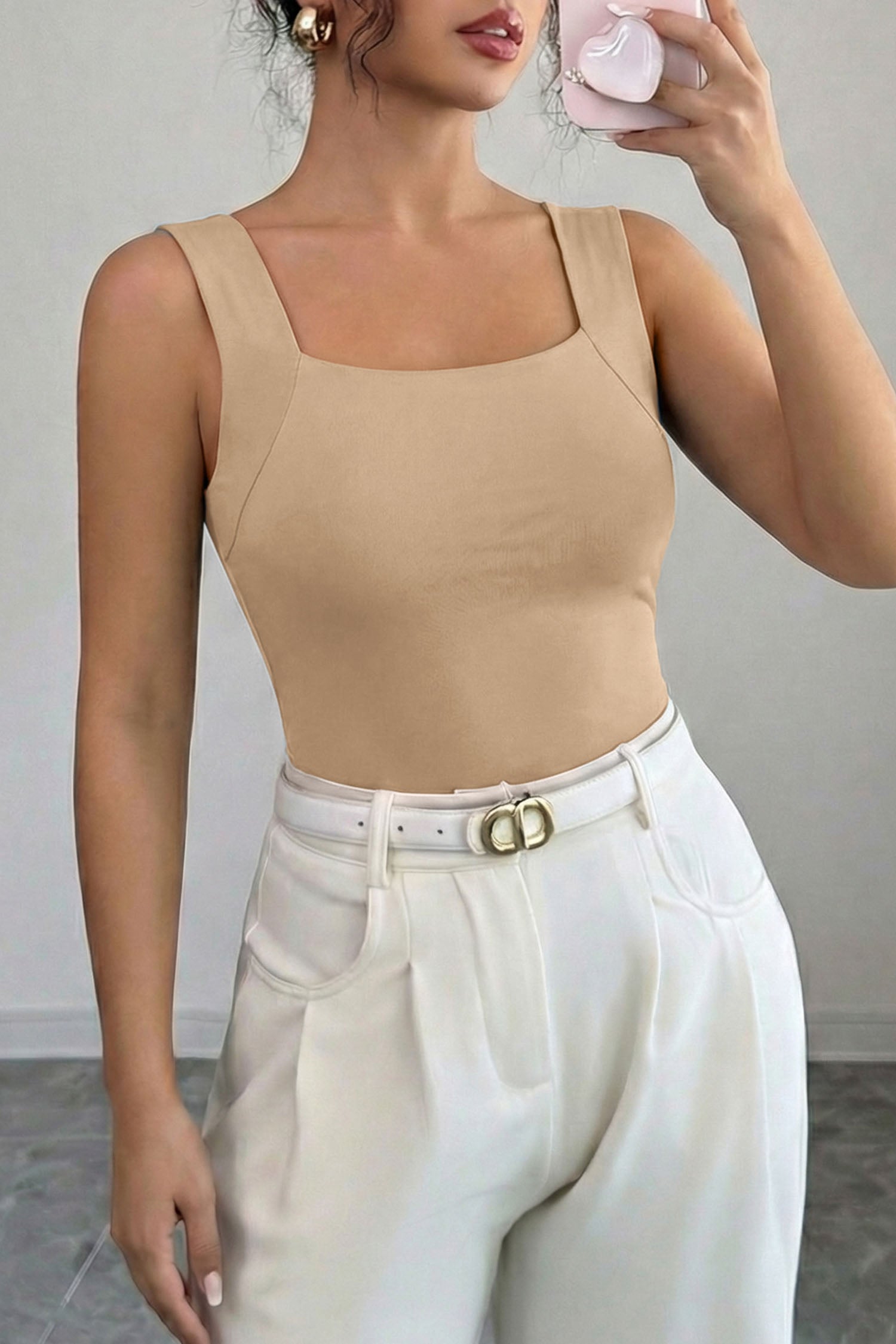 Woman wearing a beige tank top and white pants against a plain background