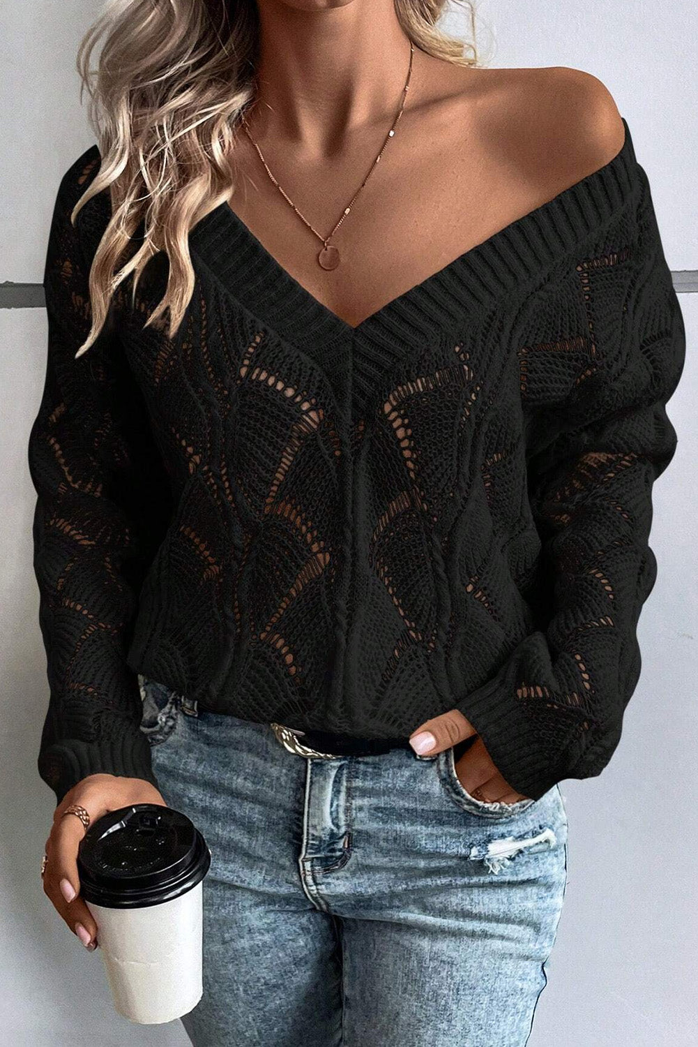 Person wearing a black knitted sweater and blue jeans, holding a coffee cup.