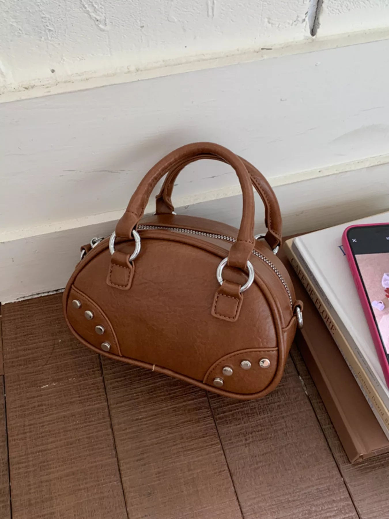 Brown handbag with silver studs on a wooden floor next to a book and phone.