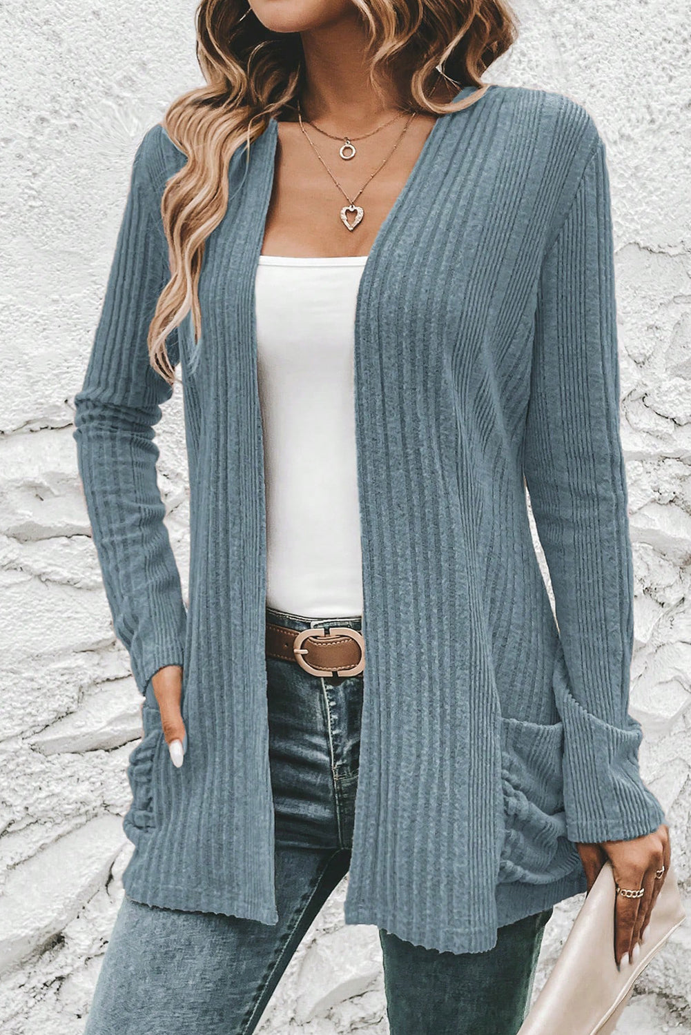 Woman wearing a blue ribbed cardigan over a white top and jeans, standing against a stone wall.