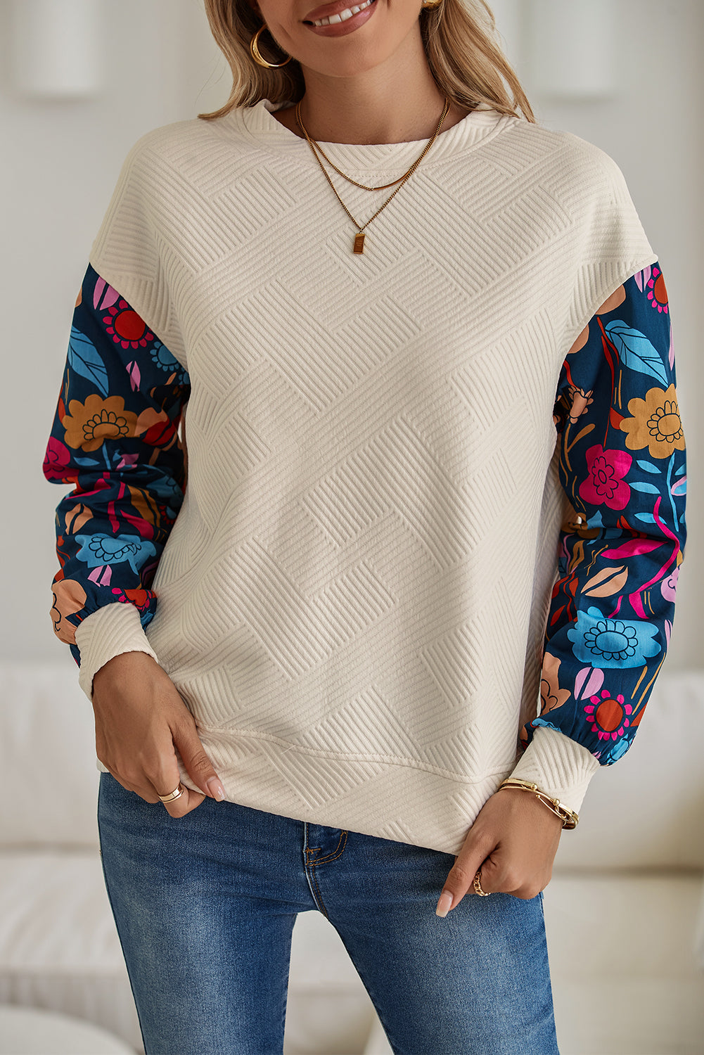Person wearing a cream sweater with colorful floral sleeves and blue jeans indoors.
