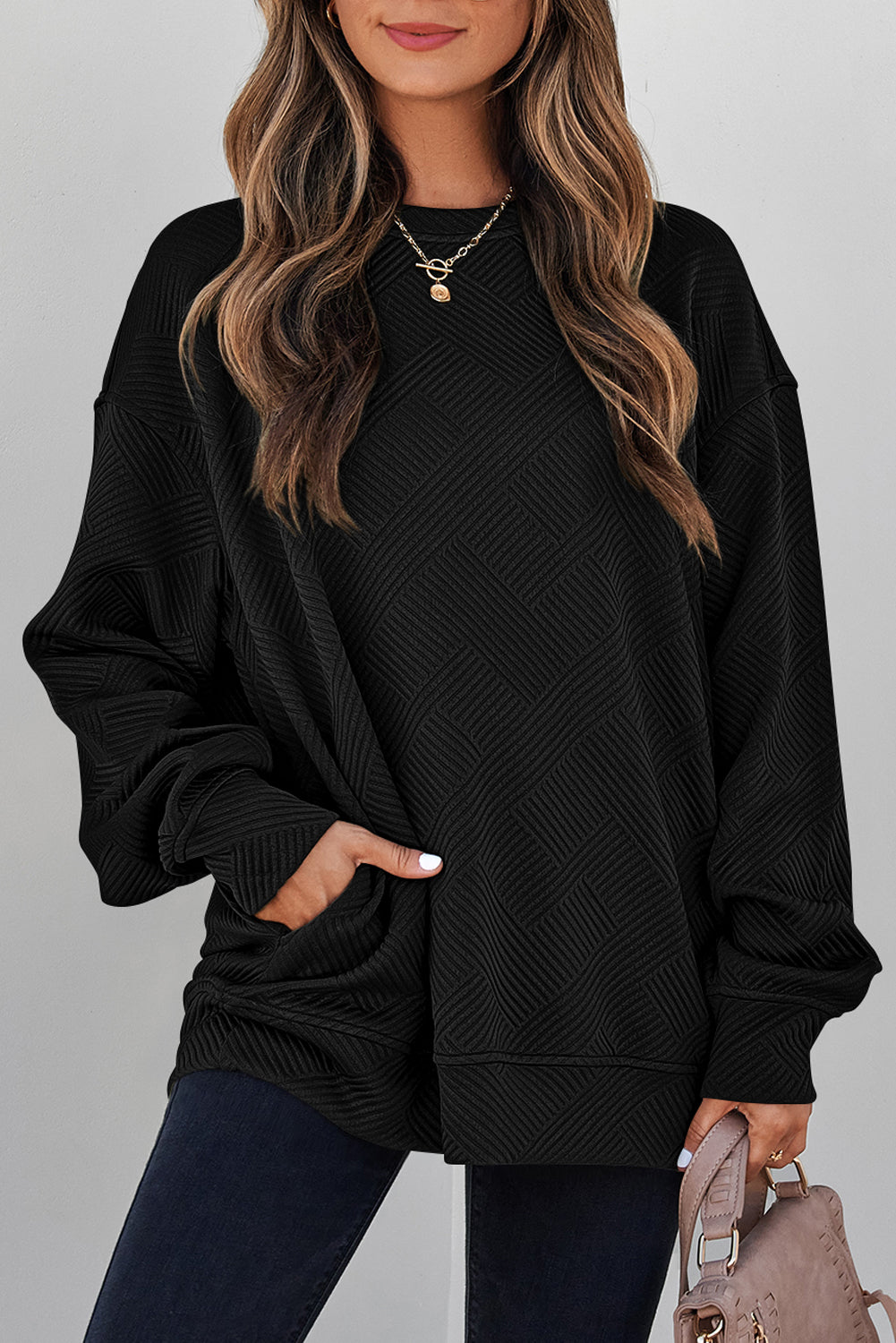 A woman wearing an oversized black Women's Textured Geometric Pullover Sweatshirt with long balloon sleeves and side pockets, paired with dark blue jeans and holding a taupe handbag against a light gray background.