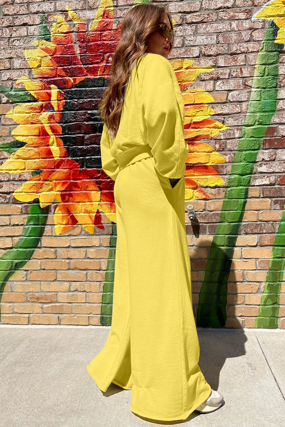 Woman in a yellow outfit standing in front of a colorful sunflower mural on a brick wall.