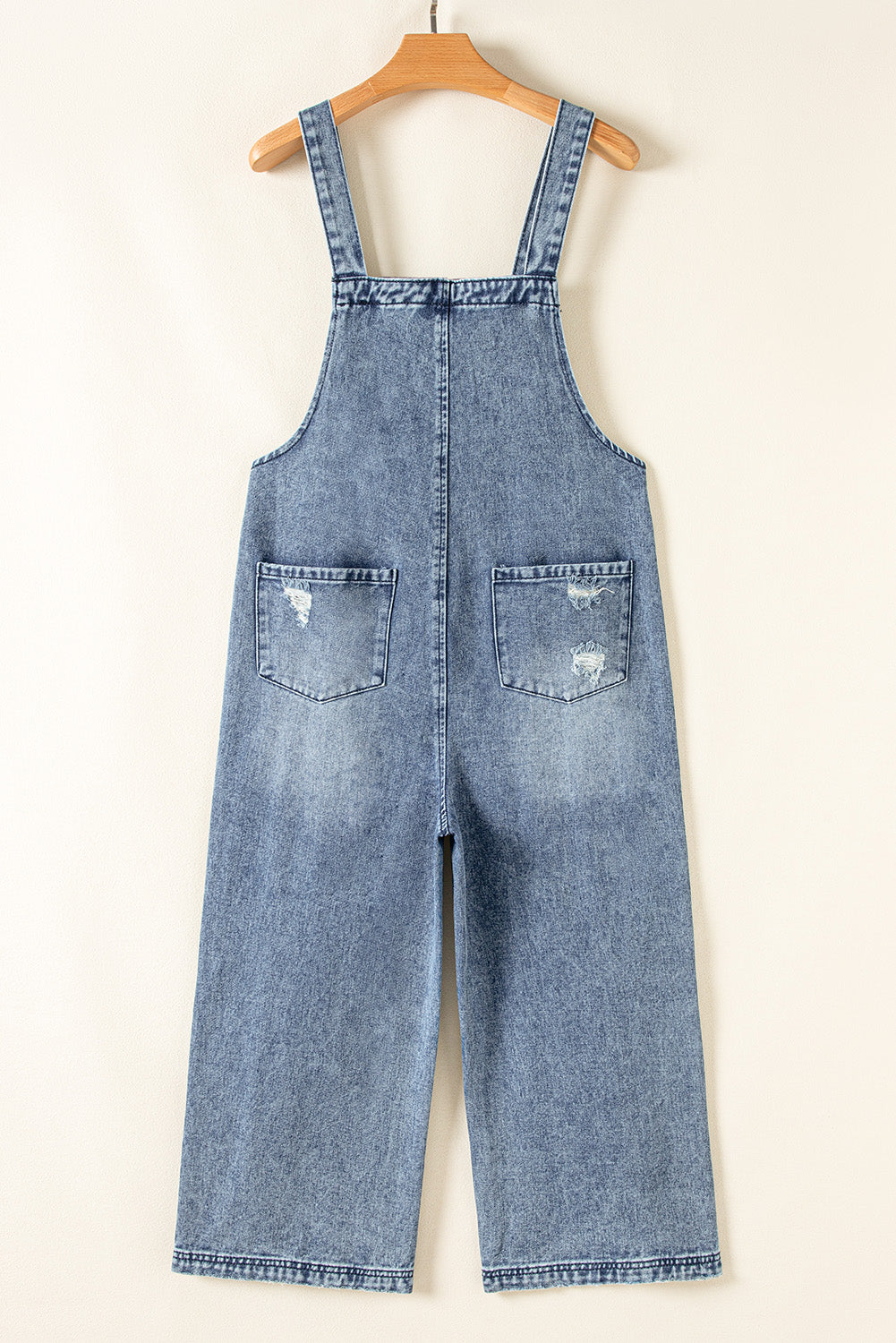Blue denim overalls on a wooden hanger against a white background