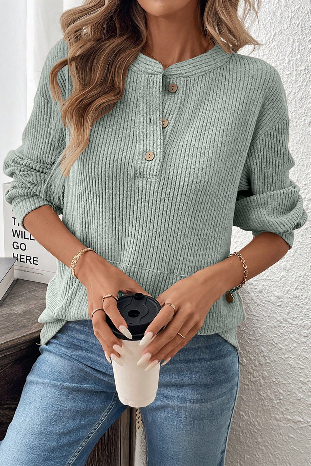 Person wearing a green ribbed sweater and blue jeans, holding a white coffee cup with a black lid.