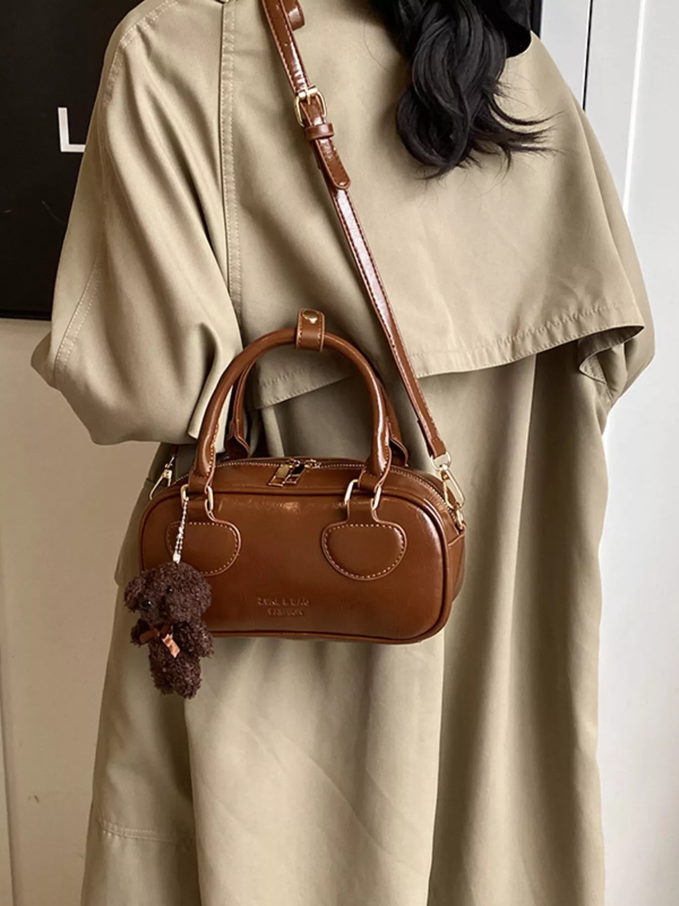 Brown handbag with a decorative charm worn by a person in a beige coat.