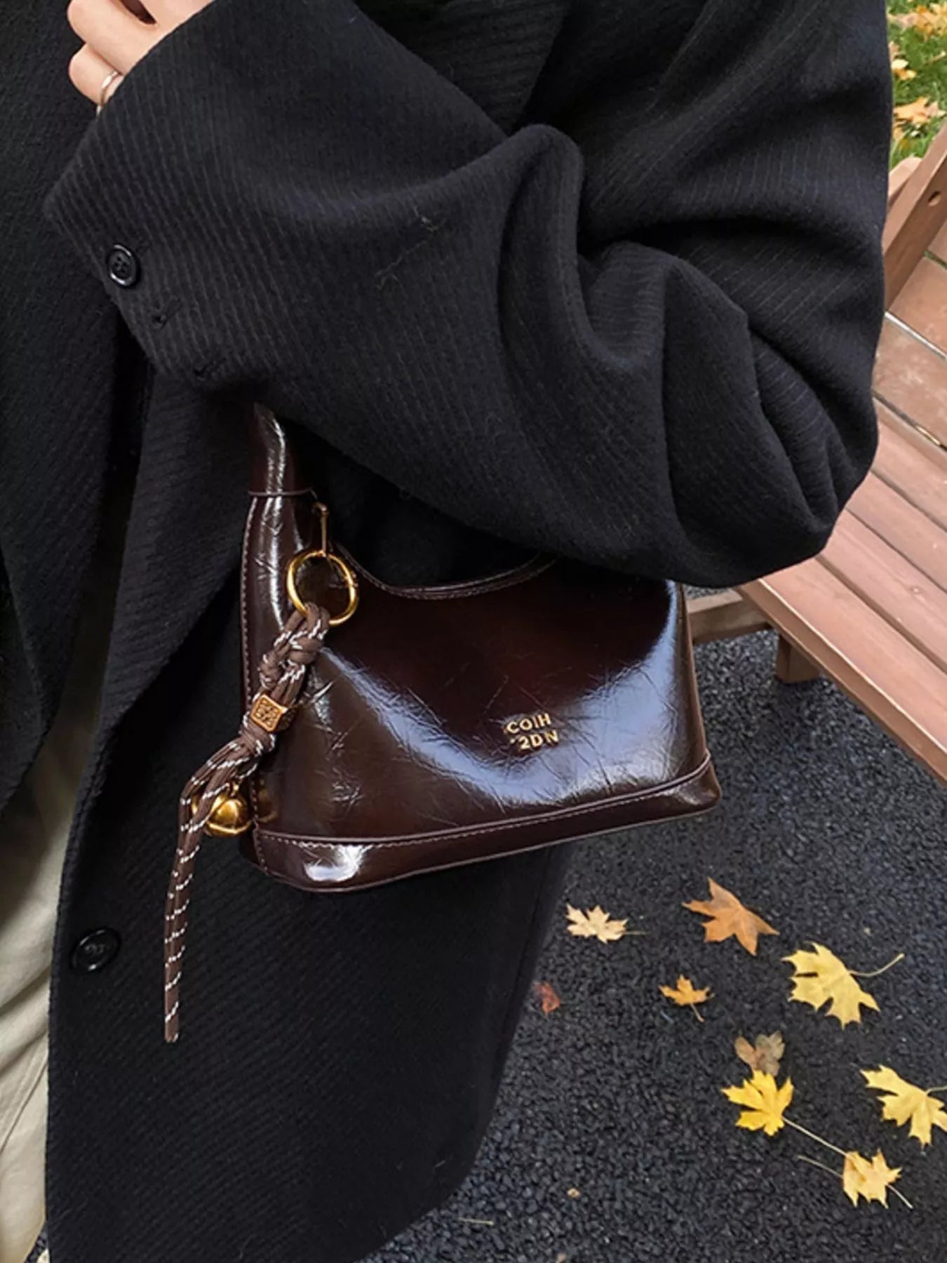 Brown leather handbag with gold chain strap held by a person wearing a black coat, with autumn leaves on the ground.