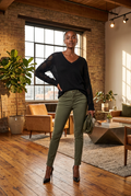 A woman wearing a black Women's Pointelle Knit V-Neck Sweater with sheer textured sleeves, olive green skinny pants, and black pointed heels. She is holding a small taupe clutch.