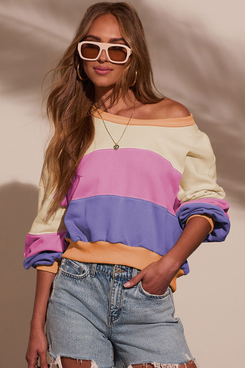 Woman wearing a colorful off-shoulder sweater and denim shorts against a neutral background