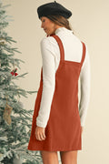 Woman wearing a rust-colored pinafore dress over a white long-sleeve shirt, standing next to a decorated Christmas tree.