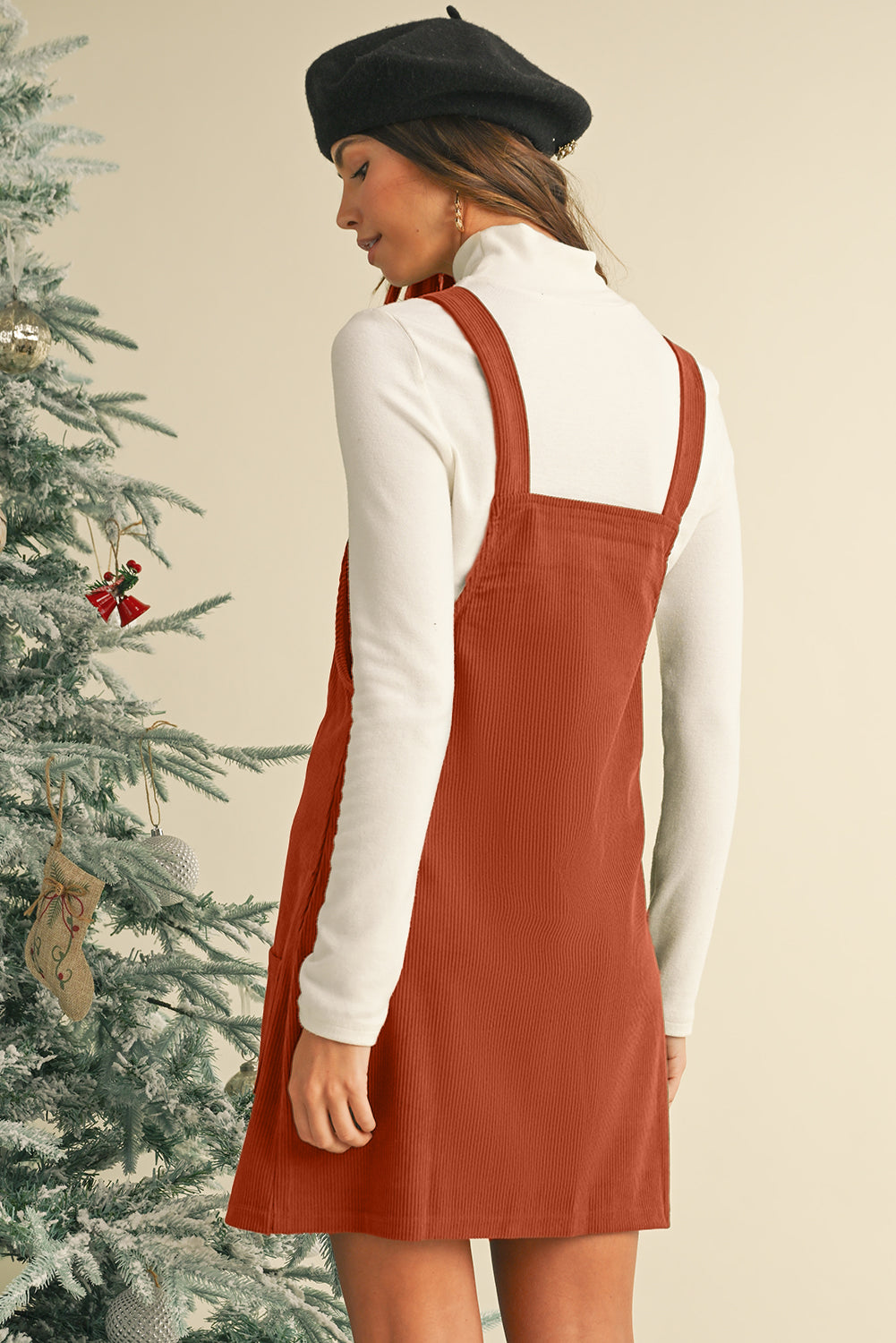 Woman wearing a rust-colored pinafore dress over a white long-sleeve shirt, standing next to a decorated Christmas tree.