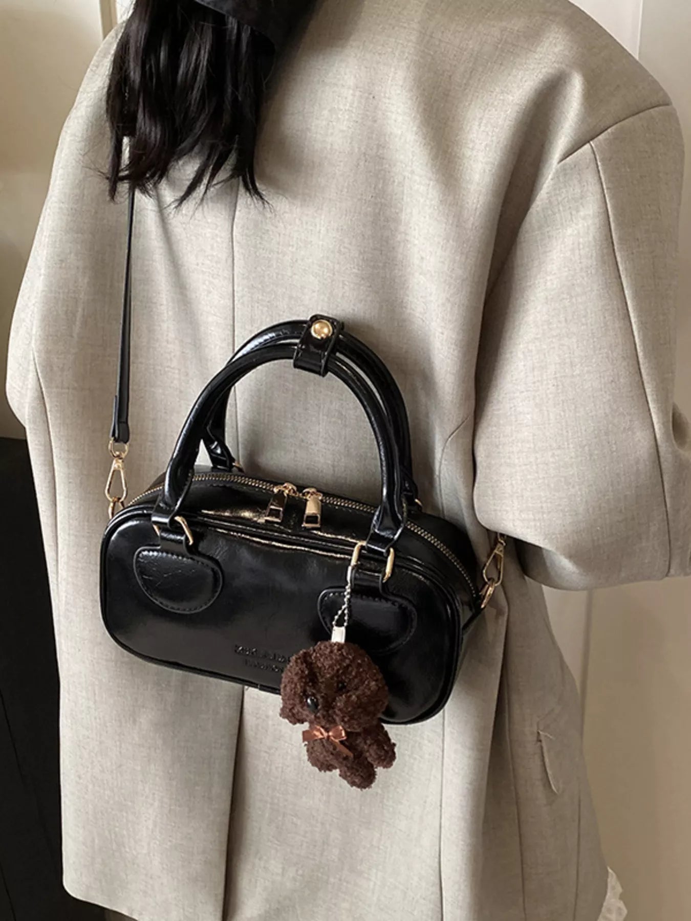 Black handbag with a decorative charm worn by a person in a beige coat.