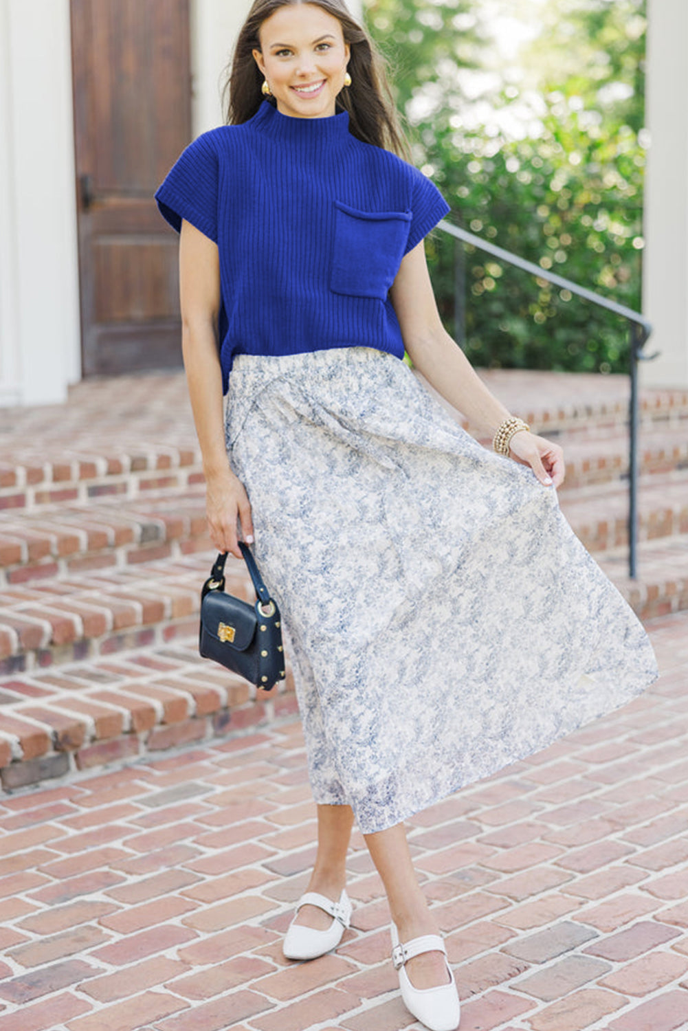 Woman wearing a blue top and floral skirt standing on a brick path with greenery in the background