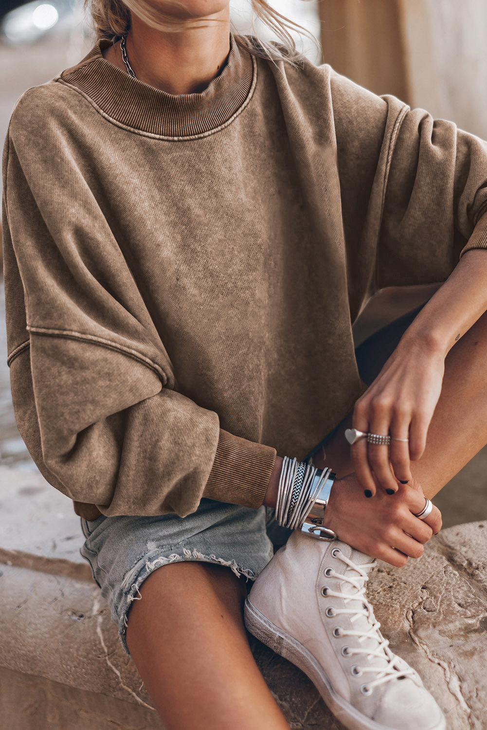 Person wearing a brown oversized sweater, denim shorts, and white sneakers sitting on a stone surface.