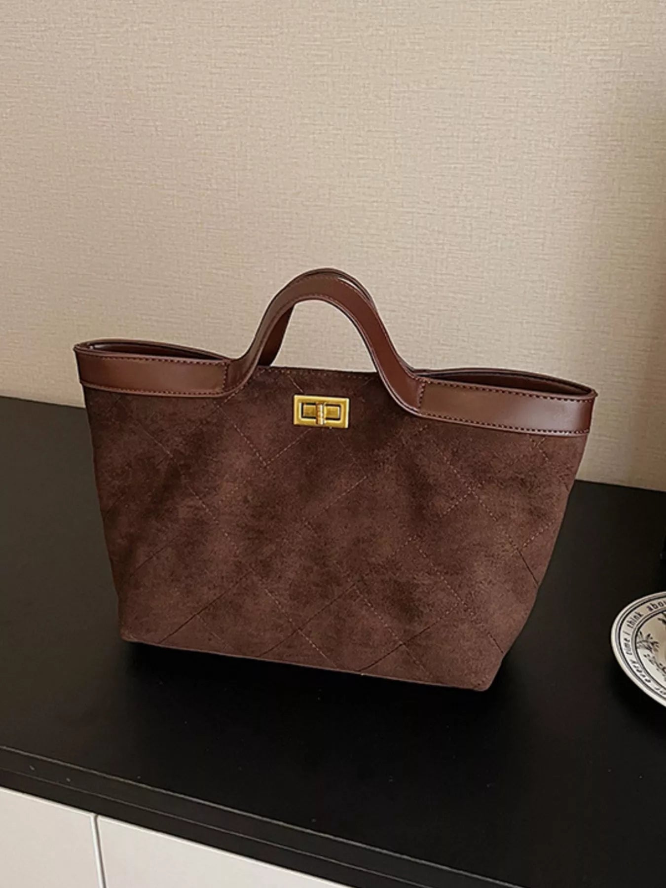 Brown handbag with a gold clasp on a dark surface against a beige wall.