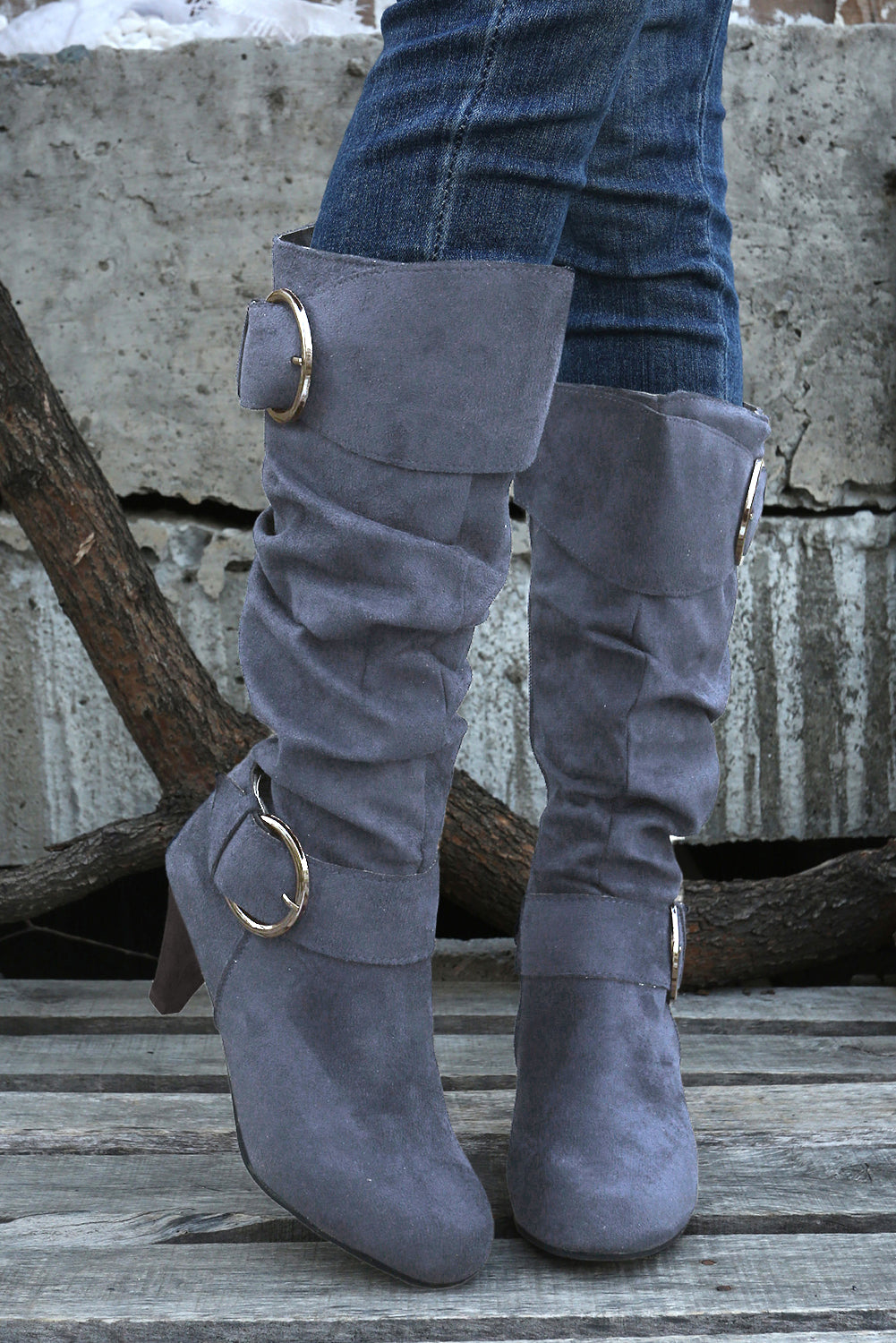 Gray suede knee-high boots with gold buckles worn with blue jeans against a rustic background.