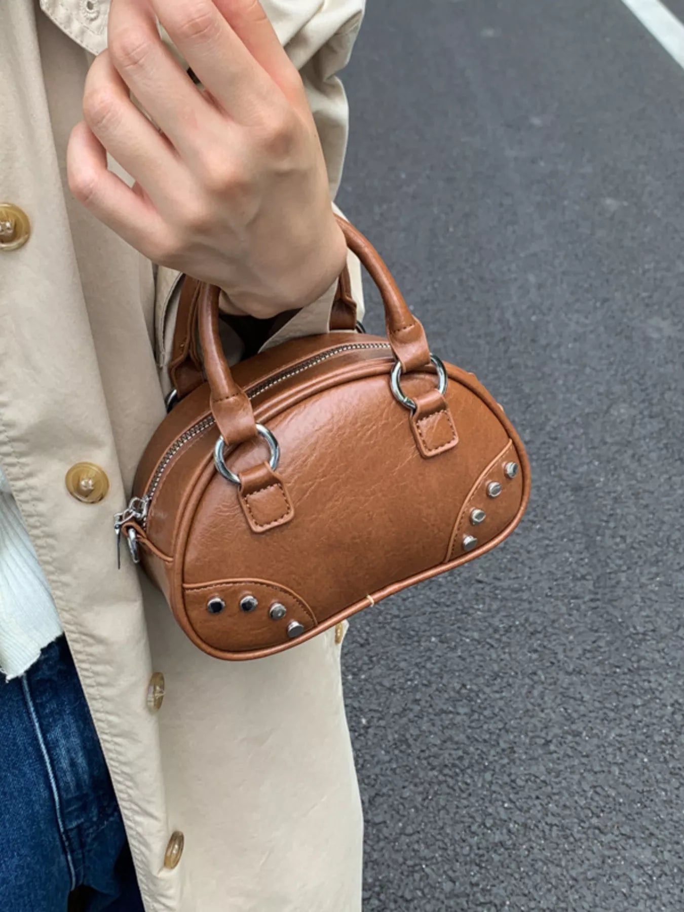 Brown handbag held by a person wearing a beige coat on a gray pavement background