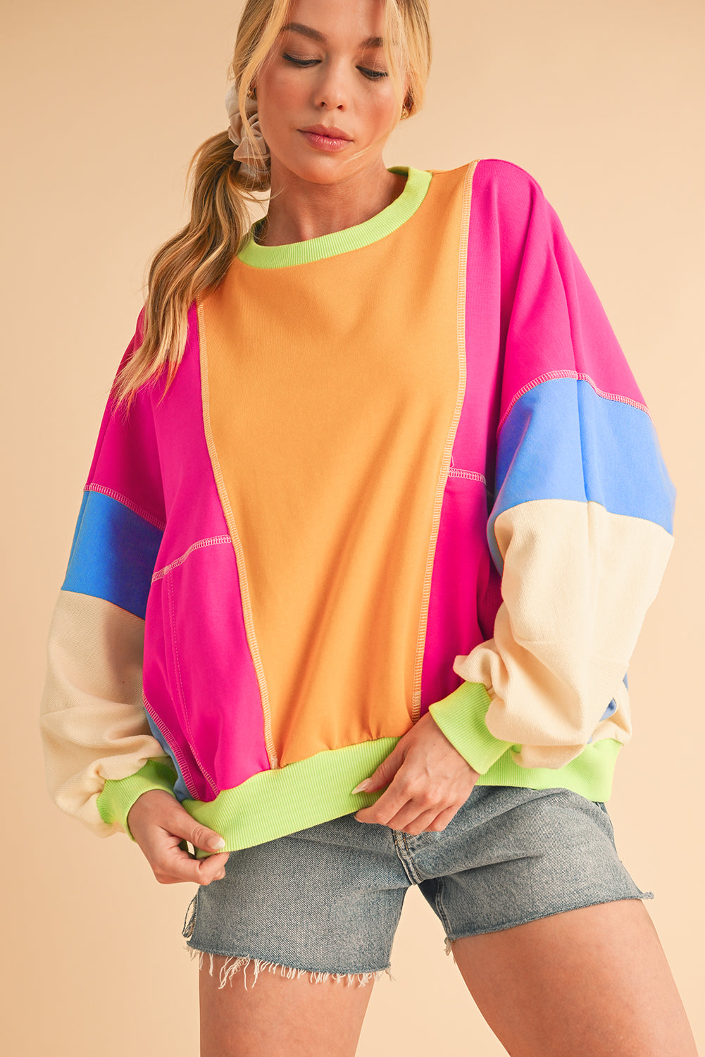 Model wearing a vibrant colorblock sweatshirt paired with denim shorts, smiling against a warm-toned backdrop