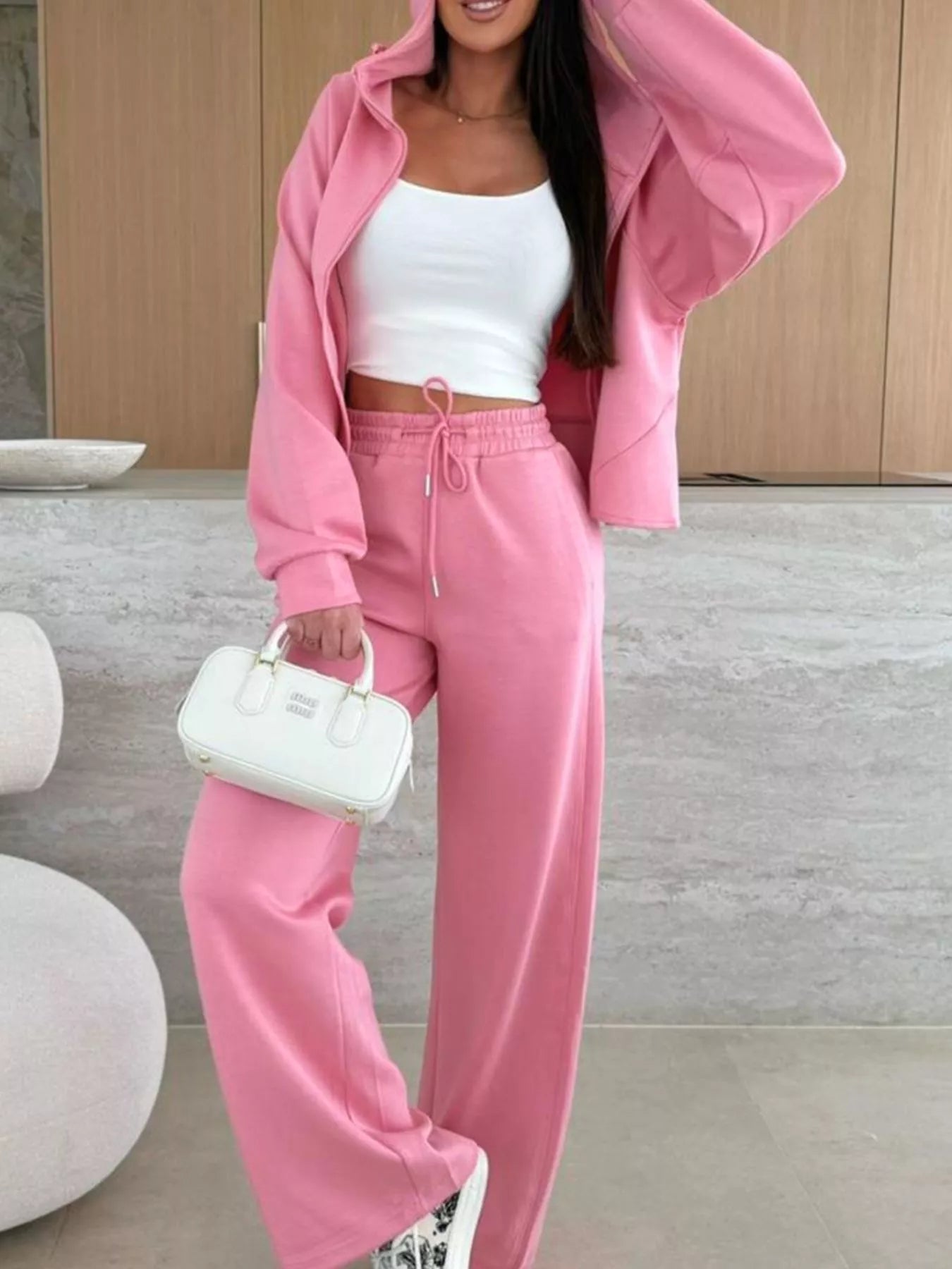 Person wearing a pink tracksuit with a white top and holding a white handbag indoors.