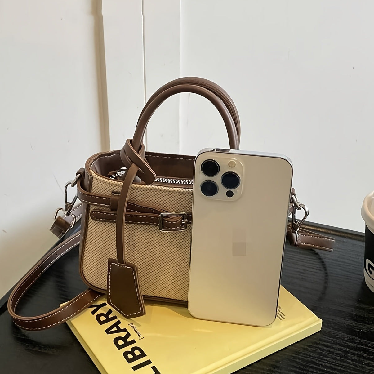 Beige handbag with brown strap, smartphone, and book on a table
