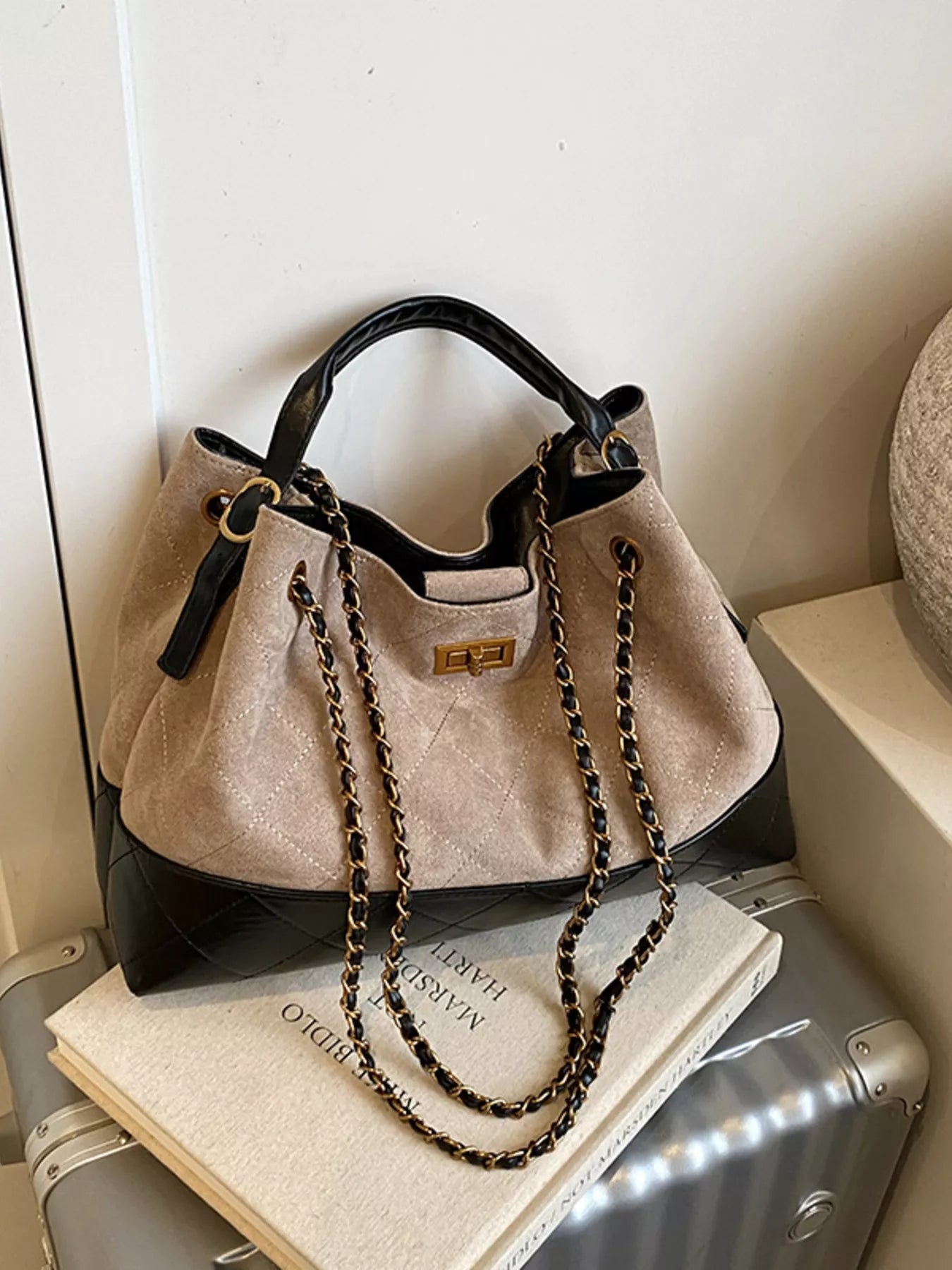 Beige handbag with black accents and a chain detail on top of a suitcase and book.