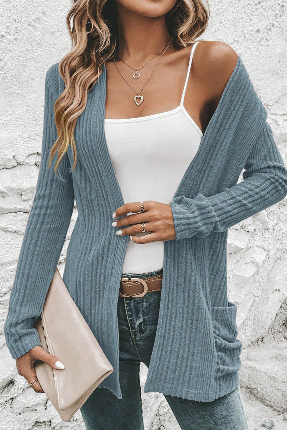 Woman wearing a blue cardigan over a white tank top with jeans and holding a beige clutch.