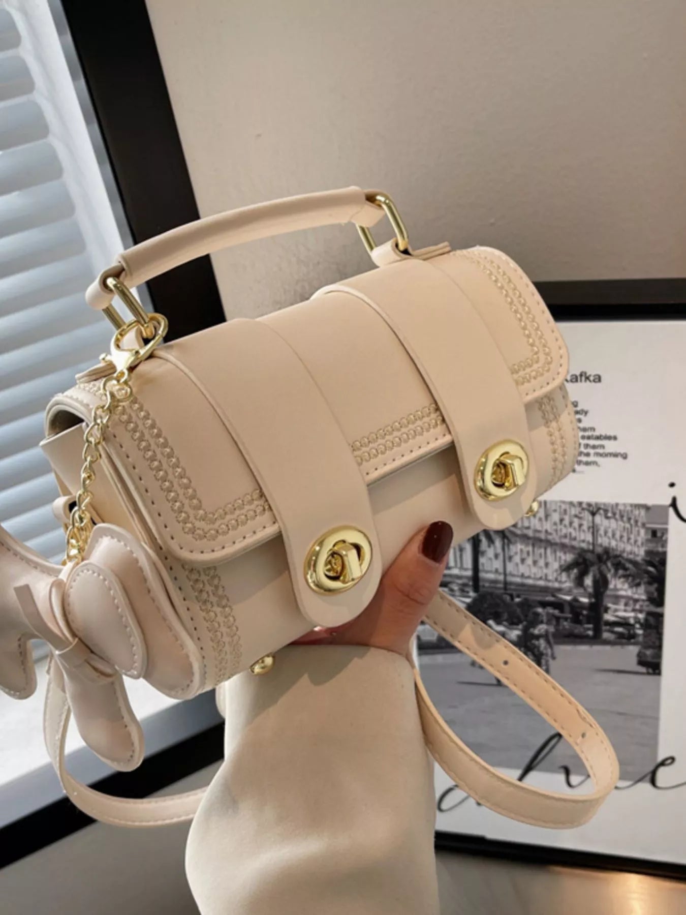 Beige handbag with gold accents held by a person in front of a window.