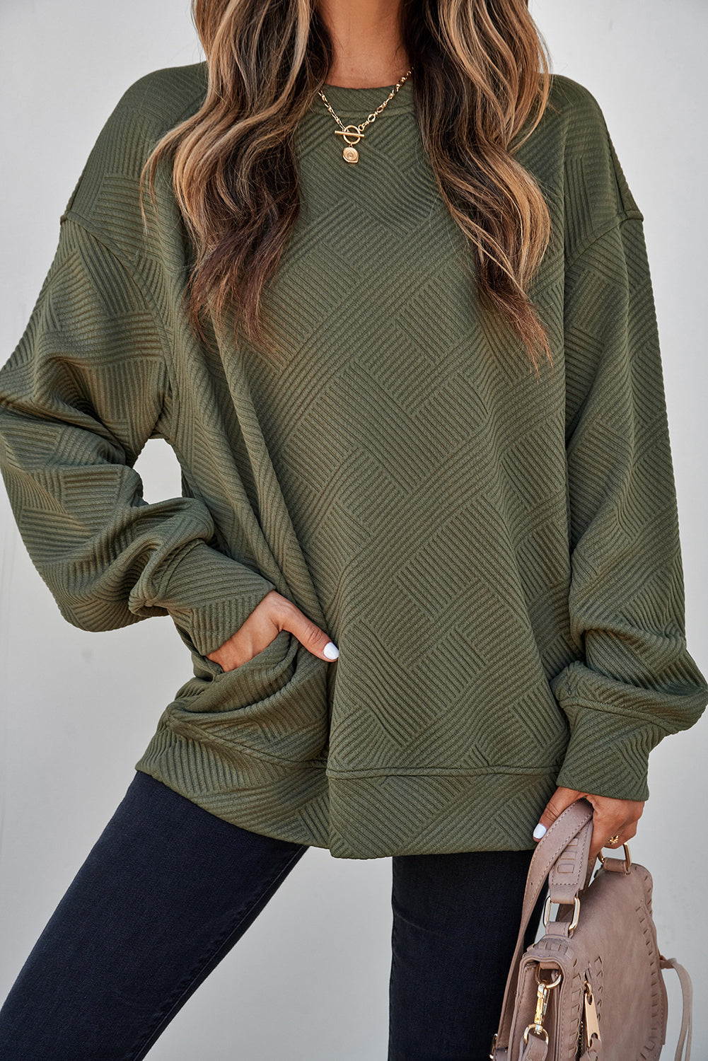 A woman wearing an olive green Women's Textured Geometric Pullover Sweatshirt with a textured chevron pattern, paired with dark jeans and holding a taupe handbag.