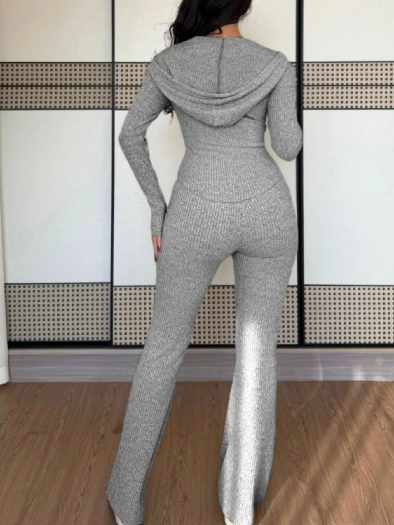 Person wearing a gray knit jumpsuit standing in front of a white wall with a geometric pattern.