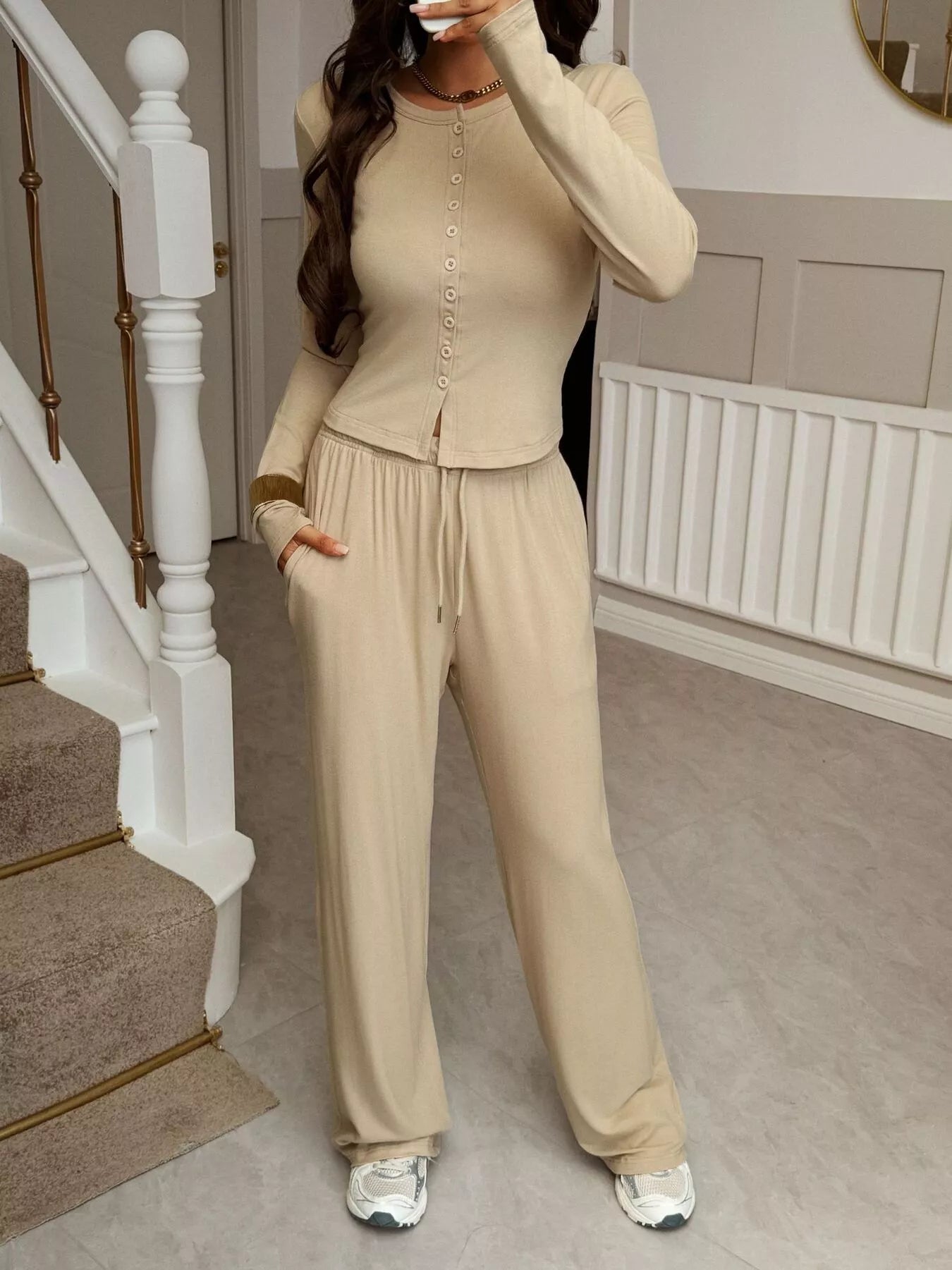 Person wearing a beige tracksuit standing in a home setting with a staircase.