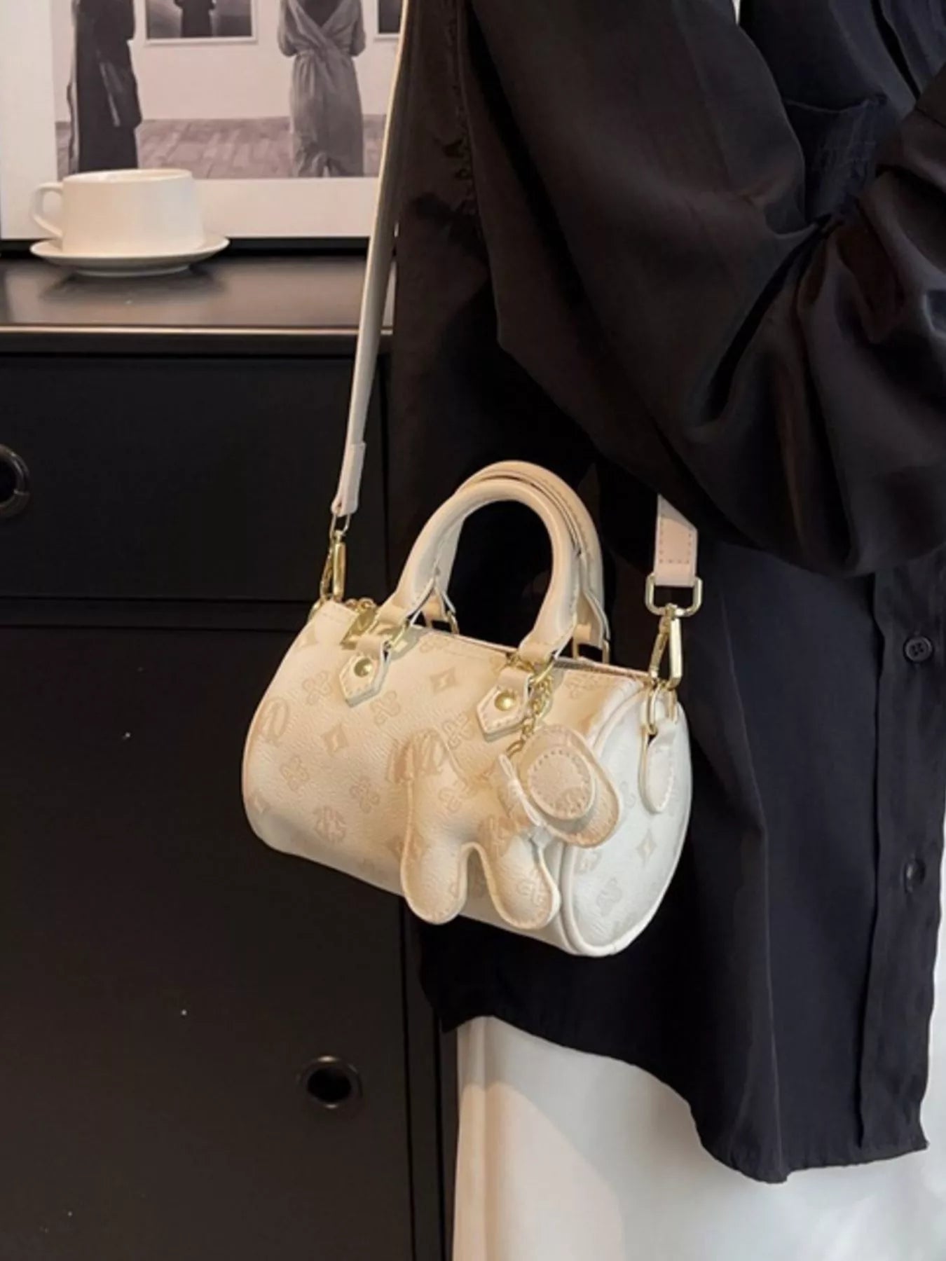 Beige handbag with gold accents hanging on a black coat against a neutral background