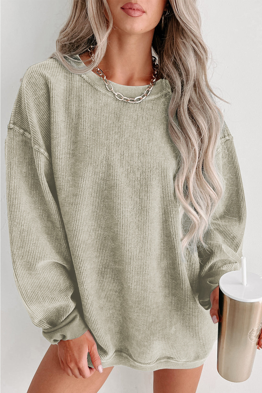 Person wearing a green oversized sweater holding a white cup with a straw on a plain background