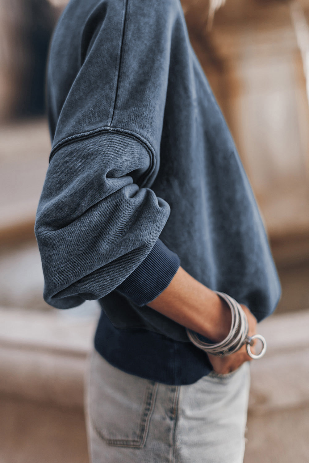 Person wearing a dark blue sweater with rolled-up sleeves, standing against a blurred background.