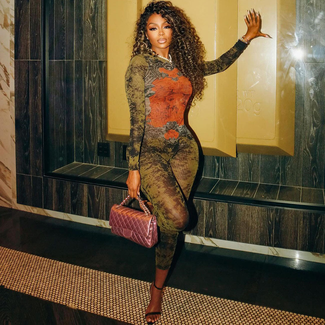 Woman in a patterned outfit holding a pink handbag in a room with a mirror.